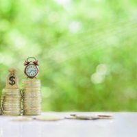 Long-tern investment, time value of money concept : Clock, US dollar bag, glass jar, a small tree on steps of rising coins. Hand pour water from a watering can, depicts asset growth from investment